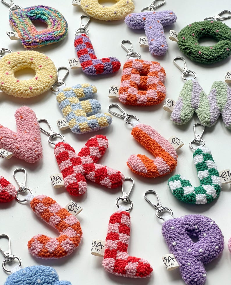 May include: A collection of colorful, handmade letter and shape keychains. Each keychain is crafted with a textured, yarn-like material and features a silver-tone clasp. The letters and shapes are in various colors and patterns, including checkerboard and polka dots.