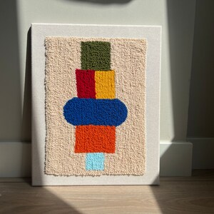 K&ouml;nnte beinhalten: Ein farbenfroher abstrakter Wandbehang mit beigefarbenem Hintergrund. Das Design zeigt einen Stapel geometrischer Formen in den Farben Gr&uuml;n, Rot, Gelb, Blau und Orange.