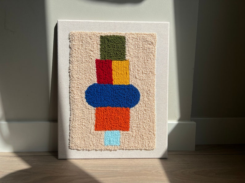 K&ouml;nnte beinhalten: Ein farbenfroher abstrakter Wandbehang mit beigefarbenem Hintergrund. Das Design zeigt einen Stapel geometrischer Formen in den Farben Gr&uuml;n, Rot, Gelb, Blau und Orange.