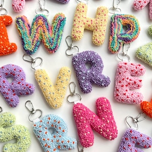 May include: A collection of colorful, handmade letter keychains. Each letter is crafted with a textured, looped yarn in various colors, including pink, purple, yellow, and blue. The keychains have silver-tone metal clasps for easy attachment.
