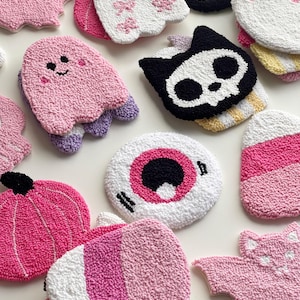 Puede incluir: Una colección de posavasos de punch needle con varias formas, incluyendo fantasmas rosas y blancos, una calavera negra y blanca, una calabaza rosa, un globo ocular blanco y rosa, dulces de maíz rosas y blancos, y murciélagos rosas texturizados.
