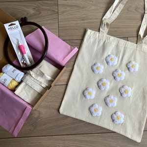 Op de afbeelding: Een witte tote bag met geborduurde witte madeliefjes. De tas zit in een doos met borduurbenodigdheden, waaronder twee garenrollen, een naald en een borduurring.