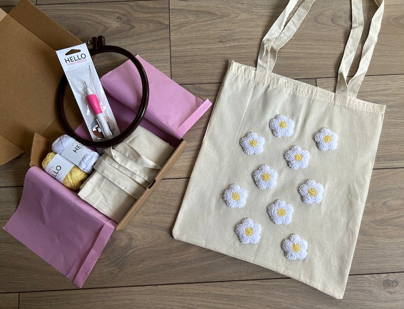 Op de afbeelding: Een witte tote bag met geborduurde witte madeliefjes. De tas zit in een doos met borduurbenodigdheden, waaronder twee garenrollen, een naald en een borduurring.