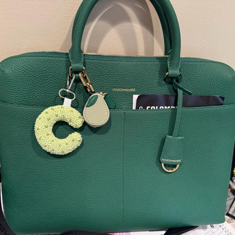 May include: A green leather handbag with a top handle and a front pocket. The bag has a gold zipper and hardware. A green crocheted letter "C" and a pear-shaped charm hang from the handle. The brand name "MAISON SABRE" is visible.
