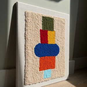 K&ouml;nnte beinhalten: Ein farbenfrohes abstraktes geometrisches Design wird mit Garn auf einer wei&szlig;en Leinwand geschaffen. Das Design zeigt gr&uuml;ne, rote, gelbe, blaue, orangefarbene und hellblaue Quadrate und Kreise.