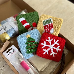Op de afbeelding: Een kerstthema punch needle-set met vijf verschillende ontwerpen: een snoepstok, een sok, een kerstboom, een sneeuwvlok en een blanco canvas. De set bevat een punch needle, garen en stof.