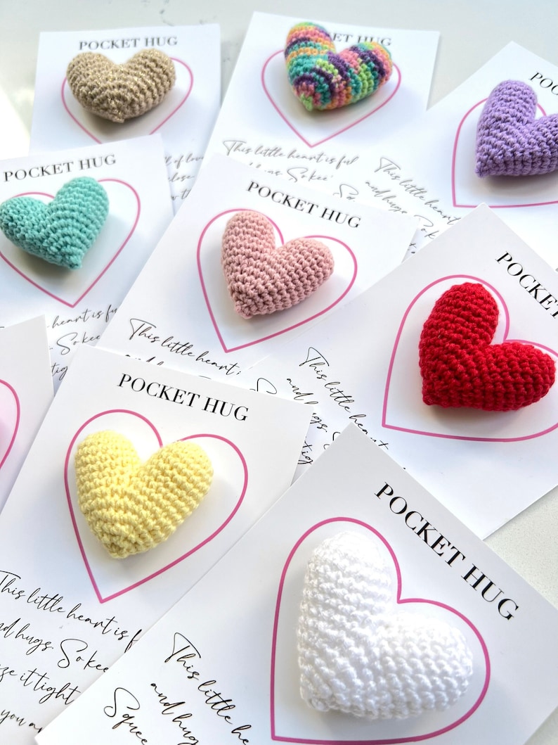 May include: Assortment of small crocheted hearts in various colors, including beige, light blue, pink, red, yellow, and white. Each heart is attached to a card with the text "Pocket Hug" and a message about the heart being a small hug.