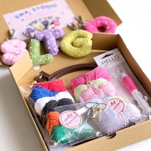 May include: A craft kit in a cardboard box, containing colorful yarn, embroidery supplies, and crochet letter keychains. The keychains spell out "LOVE" in pink, green, purple, and yellow. The kit includes a punch needle and a "HELLO" label.