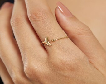 May include: A gold ring with a whale tail design. The ring is open and has a small bead at the end of the tail.