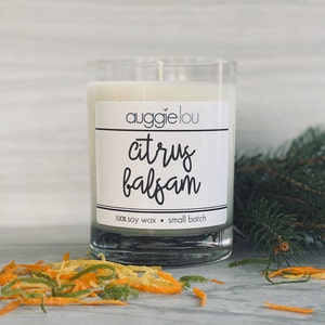 May include: A white candle in a clear glass jar with a black label that reads "auggie lou citrus balsam 100% soy wax small batch". The candle is surrounded by orange and green citrus zest.