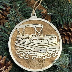 May include: A round, wooden Christmas ornament featuring a laser-cut design of a pontoon boat on water. The ornament has a silver-colored metal border and a jute string for hanging. The text "Bartlett Marina" is at the bottom.