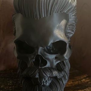 May include: A black skull sculpture with a detailed, sculpted beard and hair. The skull is made of a smooth, matte material and has a realistic appearance.