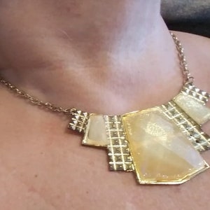 May include: A gold tone statement necklace with a geometric design. The necklace features a large, multi-faceted pendant with a yellow-gold finish.