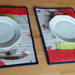 Könnte beinhalten: Zwei passende Tischsets mit weißen achteckigen Tellern, Besteck und roten und blauen Akzenten. Die Tischsets haben einen hellen Hintergrund mit einem Muster aus Erdbeeren, Pilzen und Blaubeeren. Eine grüne Serviette ist in eine Tasche gesteckt.