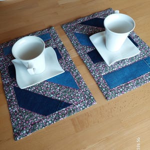 Könnte beinhalten: Zwei passende Tischsets mit floralem Muster und Jeans-Akzenten. Jedes Tischset hat eine weiße Teetasse und Untertasse. Die Tischsets liegen auf einer hellen Holzoberfläche.