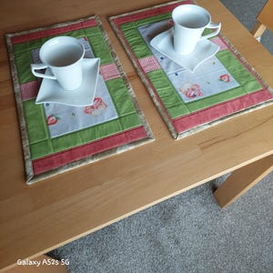 Könnte beinhalten: Zwei weiße Teetassen und Untertassen stehen auf passenden Tischsets. Die Tischsets haben ein Patchwork-Design mit grünen, roten und gemusterten Stoffquadraten. Die Tischsets haben einen Erdbeer- und Blumendruck.