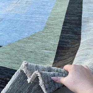 May include: A beige, brown, and blue patterned rug with a geometric design. The rug is folded over to show the backing, which is a dark gray woven fabric.