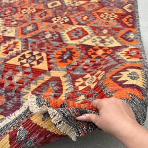 Op de afbeelding: Een kleurrijk, geometrisch patroon tapijt met een franje rand. Het tapijt is dubbelgevouwen en een hand houdt de hoek van het tapijt vast.