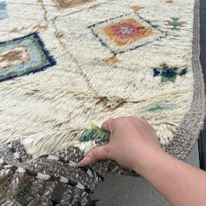 May include: Cream-colored, rectangular rug with a geometric pattern. The rug features diamond and square shapes in shades of blue, pink, orange, and brown. The edges have a gray border with tassels. The rug is made of a thick, textured material.