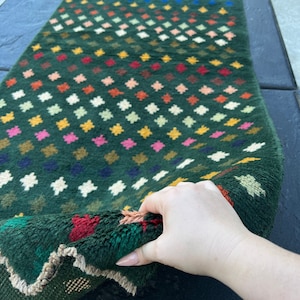 May include: A green rug with a diamond pattern in various colors, including pink, yellow, and blue. The rug is folded in half and a hand is holding the edge.