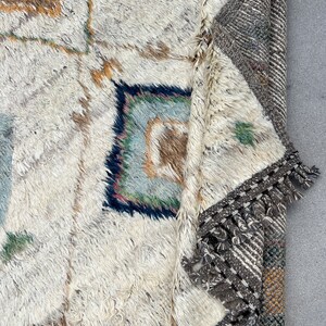 May include: A close-up of a cream-colored, textured rug with geometric patterns in shades of brown, blue, and green. The rug has a thick pile and a fringed border in gray and white. The rug is partially folded, revealing the underside.