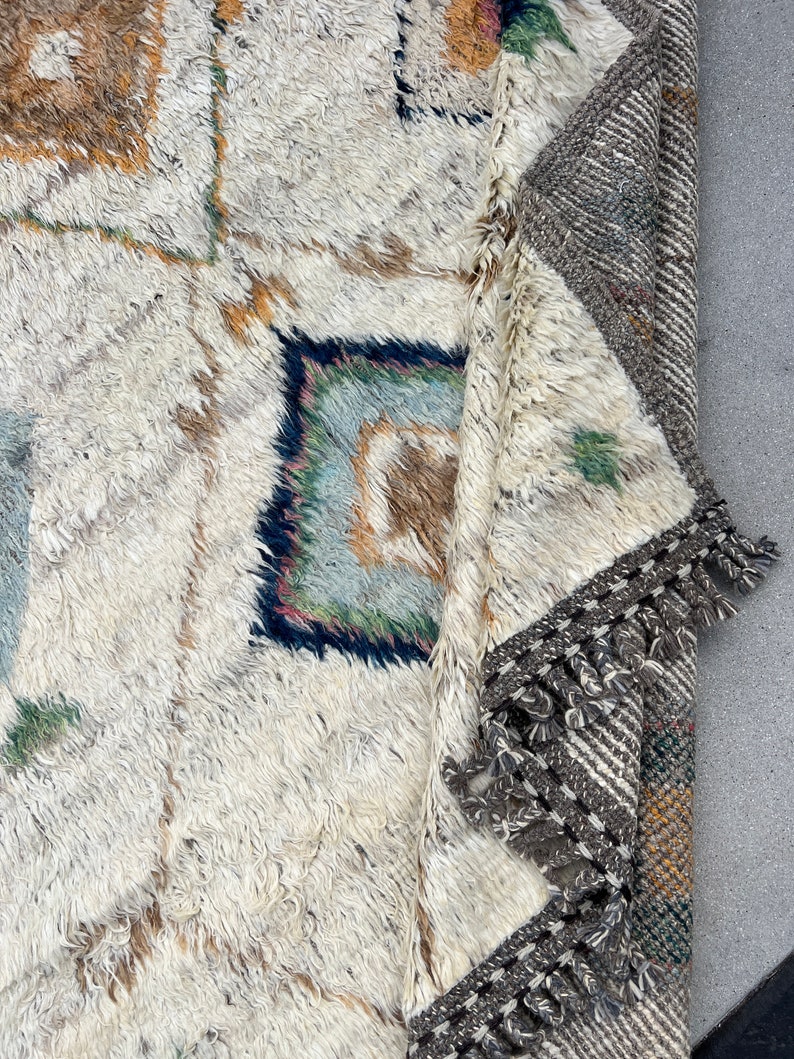 May include: A close-up of a cream-colored, textured rug with geometric patterns in shades of brown, blue, and green. The rug has a thick pile and a fringed border in gray and white. The rug is partially folded, revealing the underside.