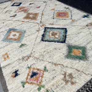 May include: Cream-colored rug with a geometric pattern of squares and diamonds in various colors, including blue, green, and pink. The rug has a textured, shaggy appearance and a fringe border.