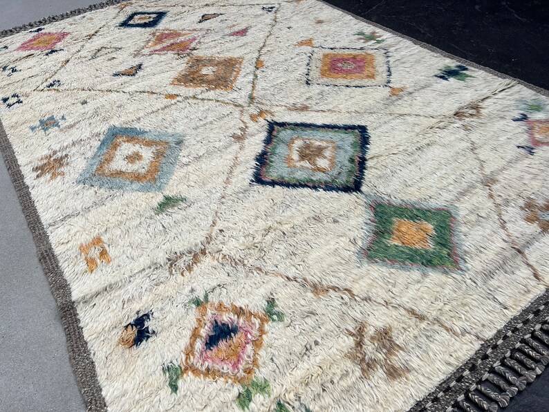 May include: Cream-colored rug with a geometric pattern of squares and diamonds in various colors, including blue, green, and pink. The rug has a textured, shaggy appearance and a fringe border.