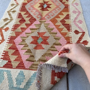 May include: A hand-woven rug with a geometric pattern in shades of cream, pink, orange, and blue. The rug features a central diamond design and fringed edges. The rug is being held up to show the underside.