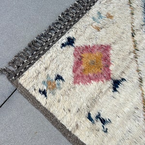 May include: A cream-colored, textured rug with a geometric design. The rug features a central pink and yellow square pattern, with scattered blue and tan accents. The border is a dark gray with fringe.