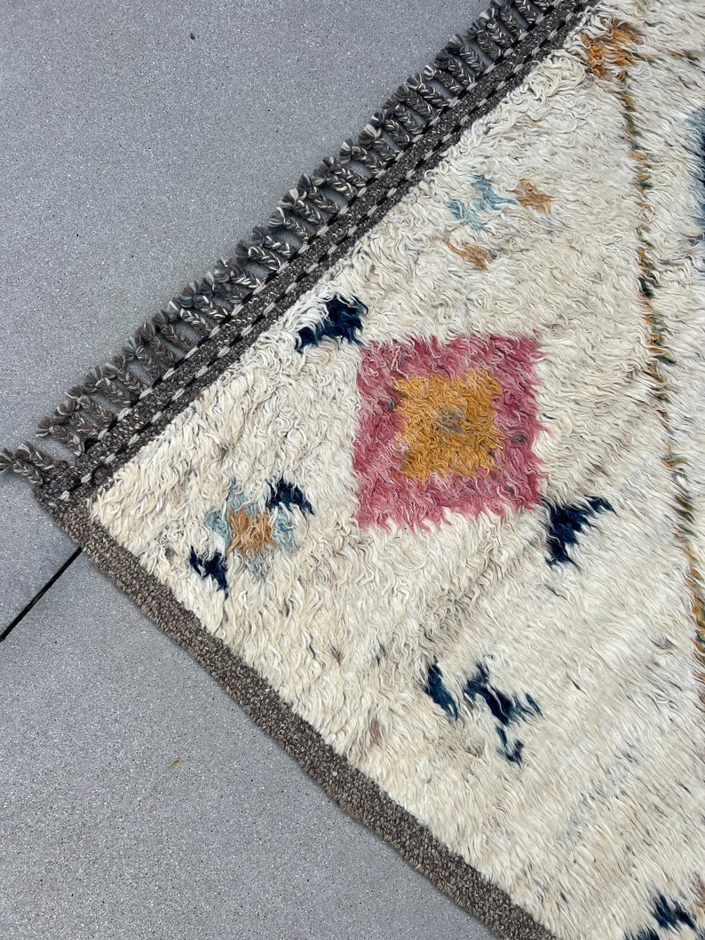 May include: A cream-colored, textured rug with a geometric design. The rug features a central pink and yellow square pattern, with scattered blue and tan accents. The border is a dark gray with fringe.