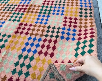 May include: A colorful, patterned rug with a peach-colored base. The rug features a repeating geometric design in squares of various colors, including blue, green, yellow, and burgundy. The rug's edge has a fringed border.