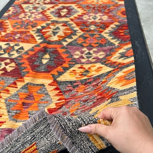 Peut inclure: Un tapis coloré avec un motif géométrique dans des tons de rouge, orange, jaune et gris. Le tapis a une bordure à franges et est plié en deux.