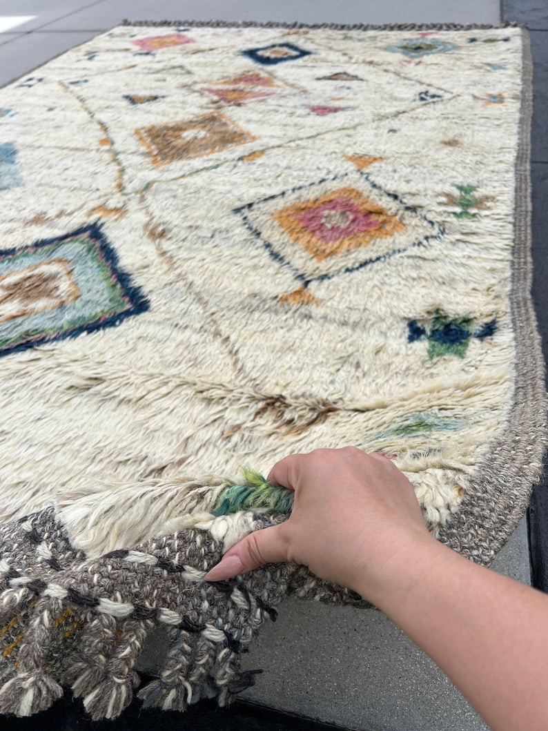 May include: Cream-colored, rectangular rug with a geometric pattern. The rug features diamond and square shapes in shades of blue, pink, orange, and brown. The edges have a gray border with tassels. The rug is made of a thick, textured material.