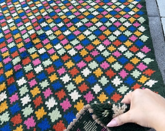 May include: A colorful, geometric patterned rug with a dark green background. The rug features a repeating diamond pattern in various colors, including blue, pink, orange, yellow, and white. The rug is made of a thick, woven material.