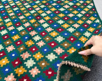 May include: A green rug with a repeating geometric pattern in various colors, including yellow, red, blue, and pink. The rug has a fringed edge and is partially folded, revealing the underside.
