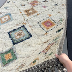 May include: Cream-colored, rectangular rug with a geometric diamond pattern in shades of blue, pink, green, and brown. The rug has a textured, shaggy appearance and a gray border with tassels along the edge. The rug is being held up.