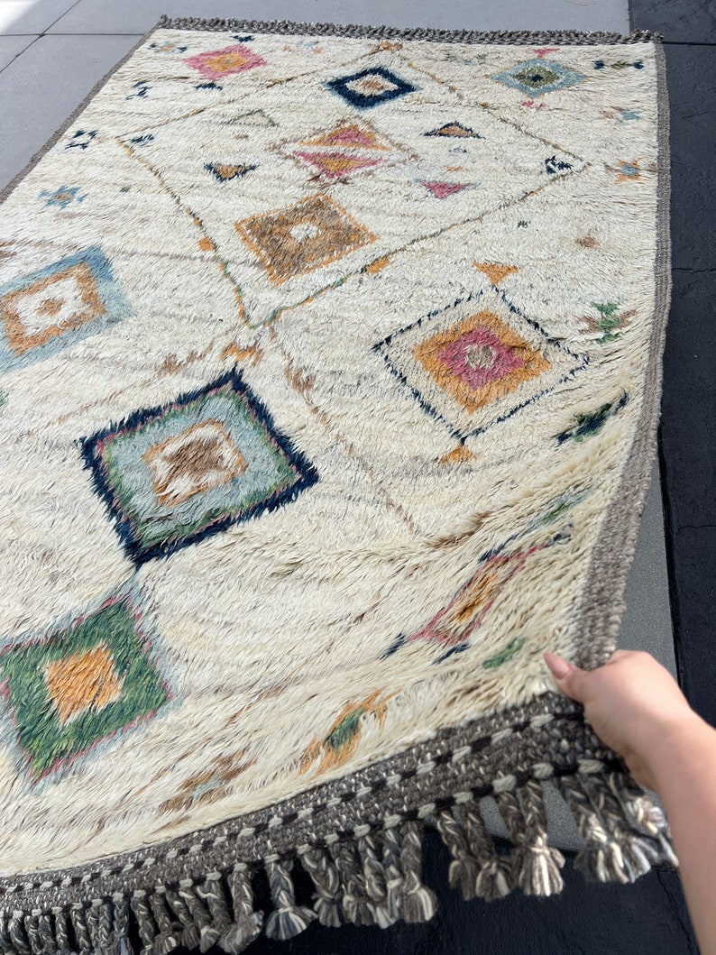 May include: Cream-colored, rectangular rug with a geometric diamond pattern in shades of blue, pink, green, and brown. The rug has a textured, shaggy appearance and a gray border with tassels along the edge. The rug is being held up.