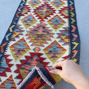 May include: A colorful, rectangular runner rug with a geometric diamond pattern in red, blue, yellow, and orange. The rug has a dark blue border and is made of woven material. The rug is being held up by a hand.