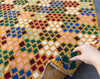 May include: A colorful, geometric patterned rug with a mustard yellow background. The rug features a repeating diamond pattern in shades of blue, green, red, pink, and white. The rug is made of a soft, textured material.