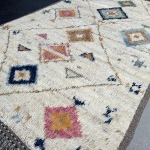 May include: Cream-colored rug with a geometric pattern. The rug features a repeating design of squares, diamonds, and triangles in shades of pink, blue, brown, and yellow. The rug has a fringed edge and a neutral border.