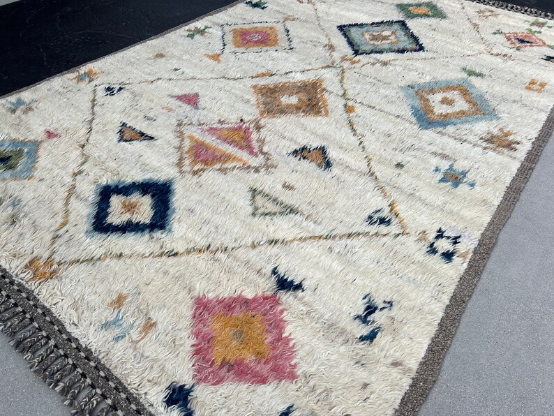 May include: Cream-colored rug with a geometric pattern. The rug features a repeating design of squares, diamonds, and triangles in shades of pink, blue, brown, and yellow. The rug has a fringed edge and a neutral border.