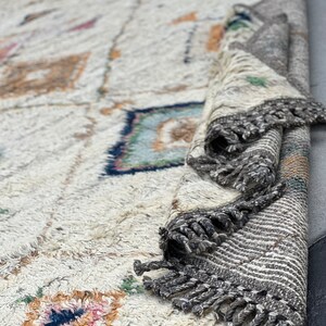 May include: Cream-colored, textured rug with geometric patterns in shades of blue, pink, and orange. The rug has a rolled-up section revealing a striped border with gray fringe. The rug's design features diamond and square shapes.