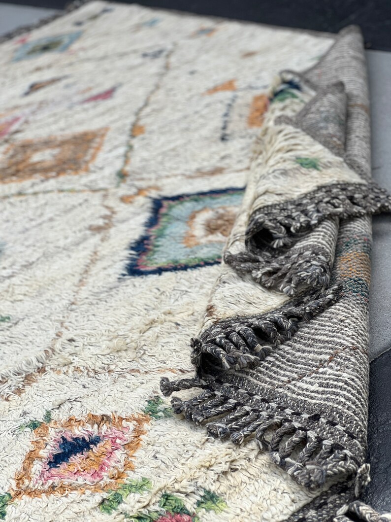 May include: Cream-colored, textured rug with geometric patterns in shades of blue, pink, and orange. The rug has a rolled-up section revealing a striped border with gray fringe. The rug's design features diamond and square shapes.