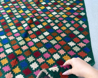 May include: A hand-woven, rectangular rug with a dark green background and a repeating pattern of colorful, stylized floral shapes. The shapes are in shades of red, orange, yellow, pink, blue, and white. The rug's edge is trimmed with a beige fringe.