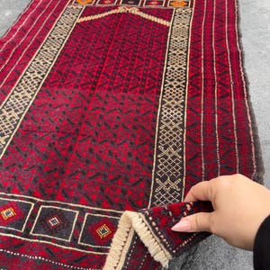 Puede incluir: Una alfombra de oración roja y negra con intrincados patrones geométricos y una franja de color crema. La alfombra tiene un diseño tradicional con un arco central y elementos decorativos.