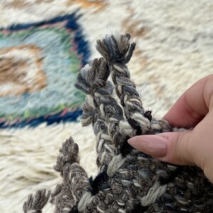 May include: Close-up of a section of a woven rug with a textured, neutral color palette. The rug features a mix of gray, black, and white yarns, with a braided fringe detail. The background shows a portion of the rug with a blue and green design.