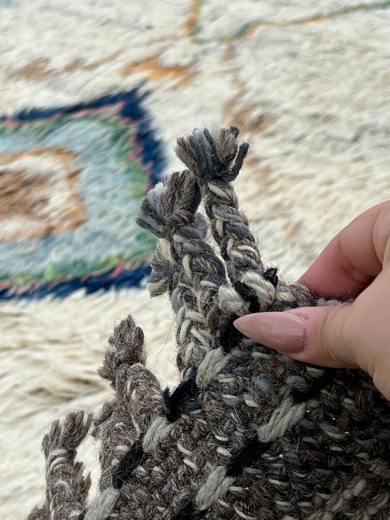 May include: Close-up of a section of a woven rug with a textured, neutral color palette. The rug features a mix of gray, black, and white yarns, with a braided fringe detail. The background shows a portion of the rug with a blue and green design.