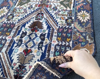 Puede incluir: Una alfombra turca vintage con un patrón geométrico en tonos azules, marrones, blancos y rojos. La alfombra tiene un borde con flecos y está doblada por la mitad.