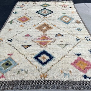 May include: A rectangular, off-white rug with a geometric diamond pattern. The design incorporates various colors, including blue, pink, yellow, and brown. The rug has a textured, shaggy appearance and a fringe border. The rug's dimensions are not visible.